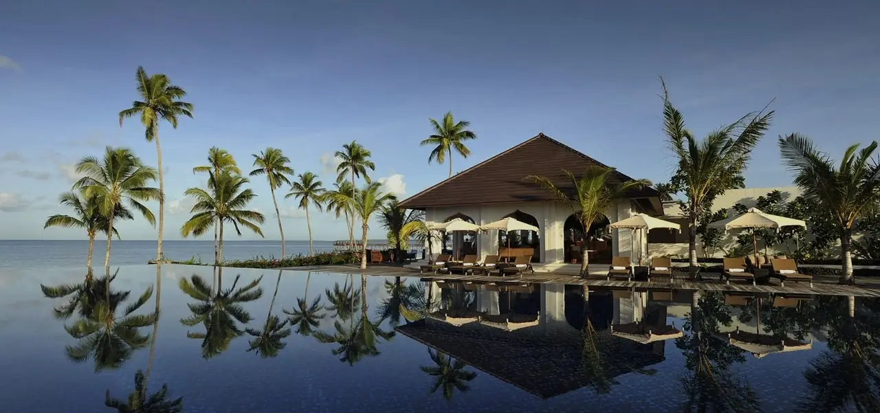 A long infinity pool reflecting palms and a thatched pavilion under a blue sky