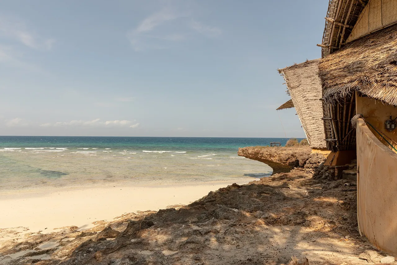 A sandy beach with clear water and a thatched building edge on the right