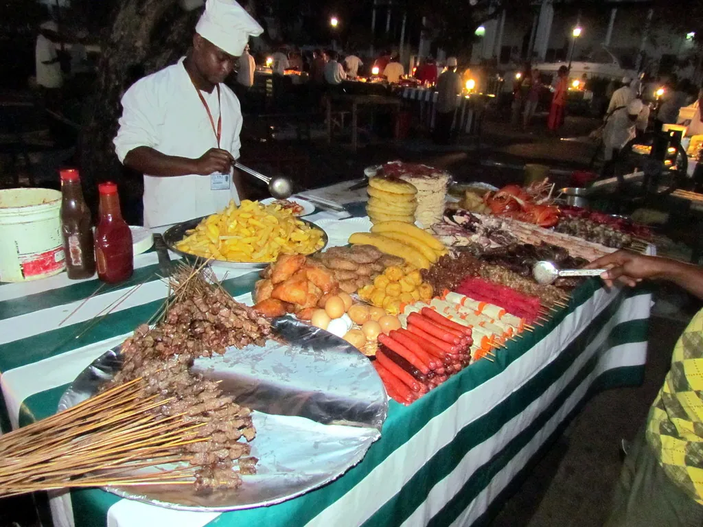 A night-market stall piled with skewers of seafood, sausages and other grilled food, with a cook in a chef's hat