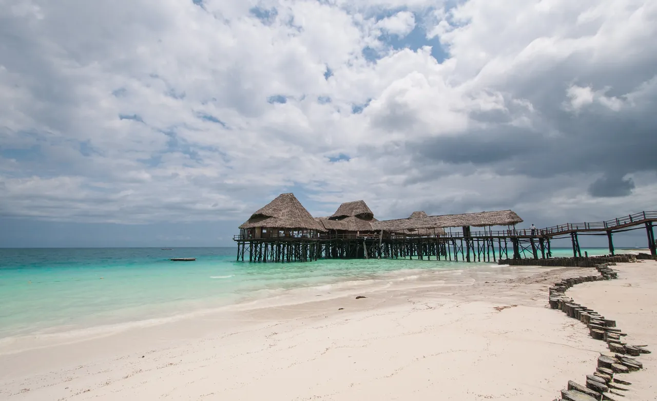 Thatched restaurant on stilts at the end of a long wooden pier over turquoise water, white sand in the foreground