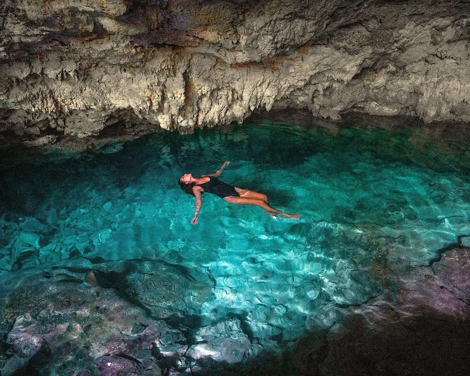 A swimmer floating on her back in clear turquoise water inside a cave with a rocky ceiling.