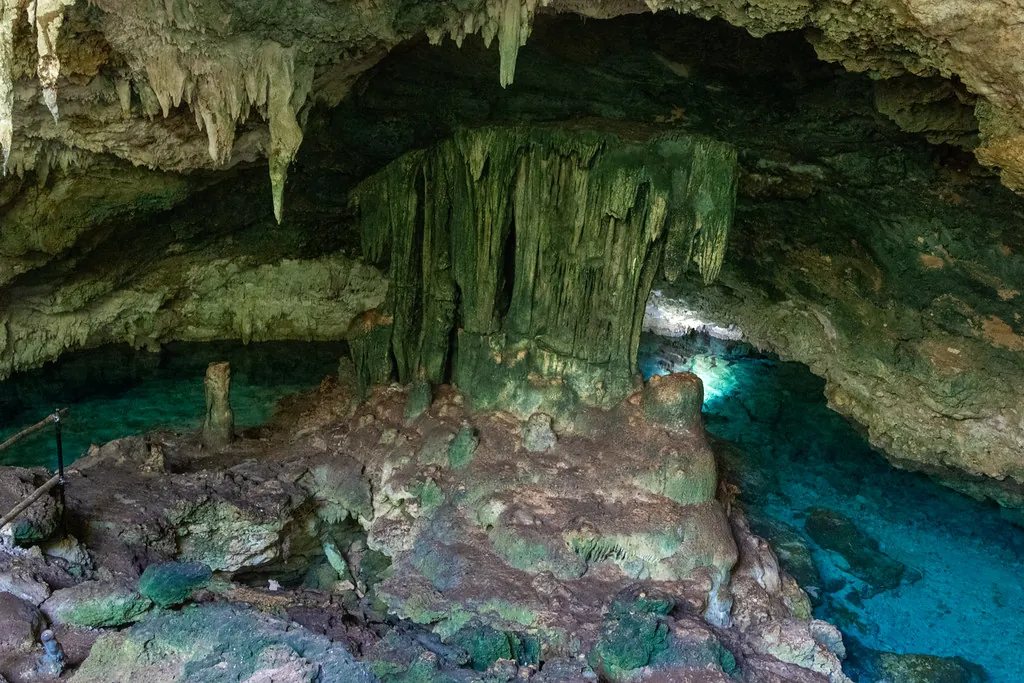 Inside a limestone cave: stalactites and a thick rock column above a pool of clear blue water.