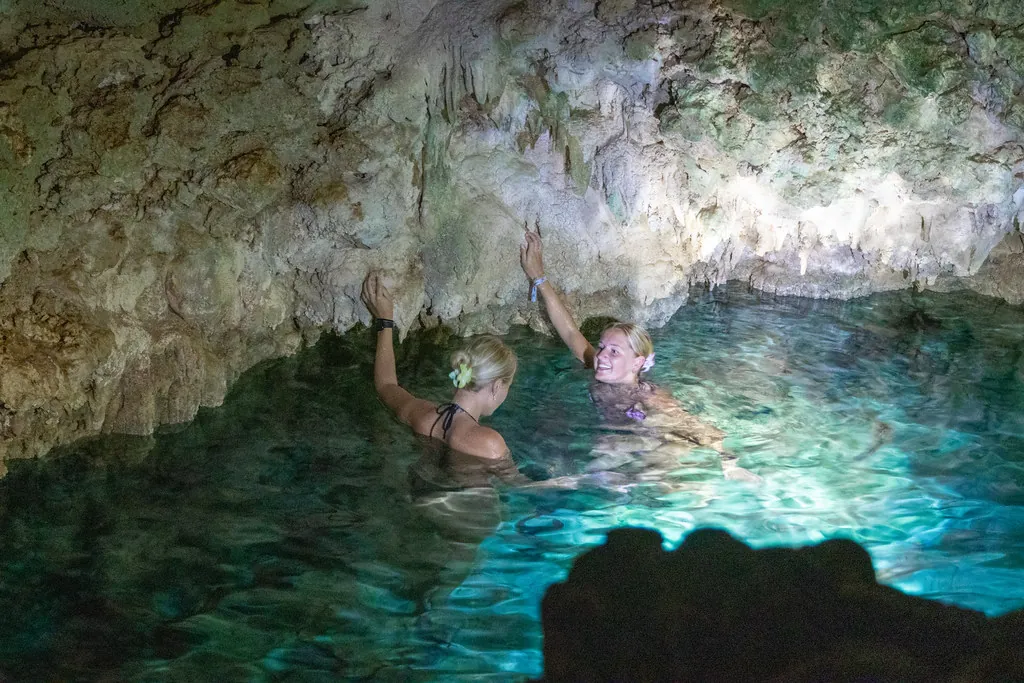Two swimmers reaching up towards the cave ceiling while standing in glowing blue water.