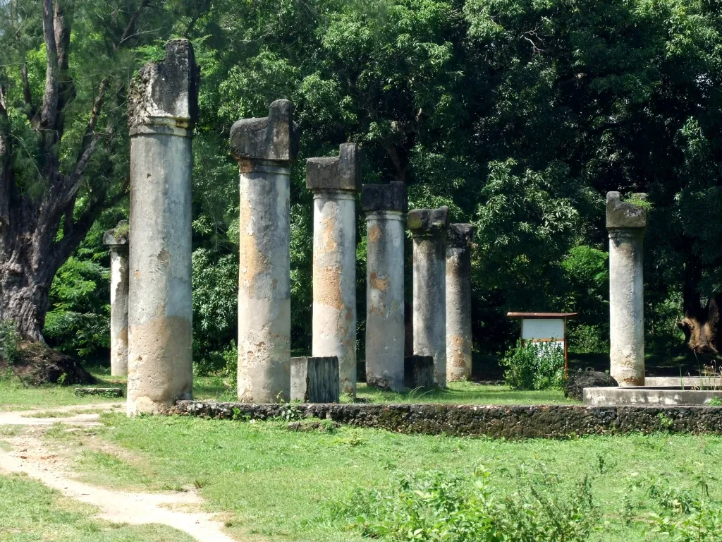 A row of tall weathered stone columns standing on grass in front of dense trees at the Maruhubi ruins