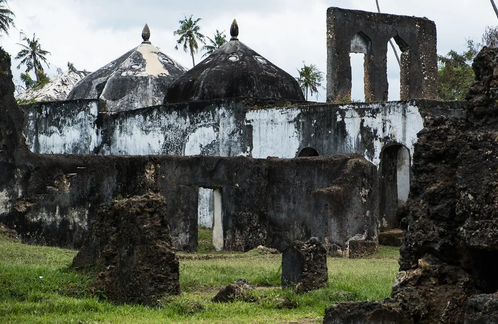 The ruined bath complex with small domed roofs and an arched wall, surrounded by grass and palms
