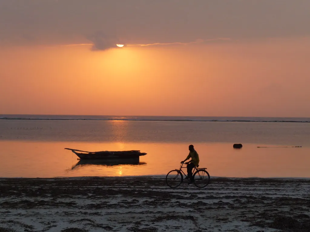 Sunrise over the sea with a cyclist silhouetted on the wet sand and an outrigger boat in the shallows