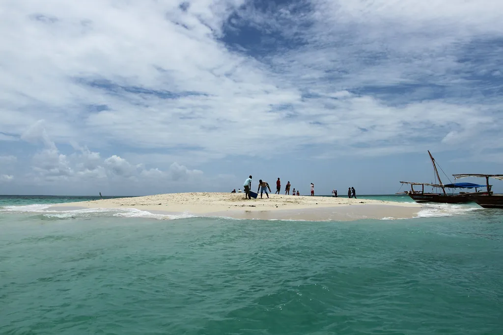 A white sandbank in green-blue water with a few people standing on it and a dhow anchored alongside.