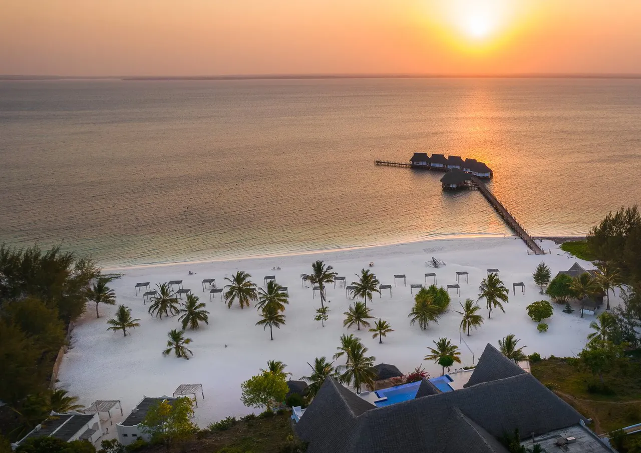 Aerial view at sunset of a palm-dotted white beach on a calm bay, with a long jetty