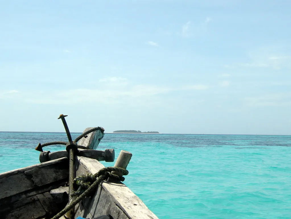 Bow of a wooden boat with an anchor heading across turquoise shallows towards a distant low island