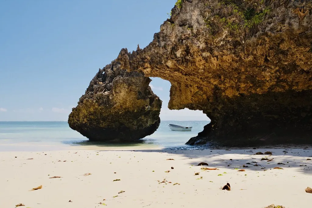 A large weathered coral-rock arch on a white-sand beach, with calm pale-blue sea behind it.