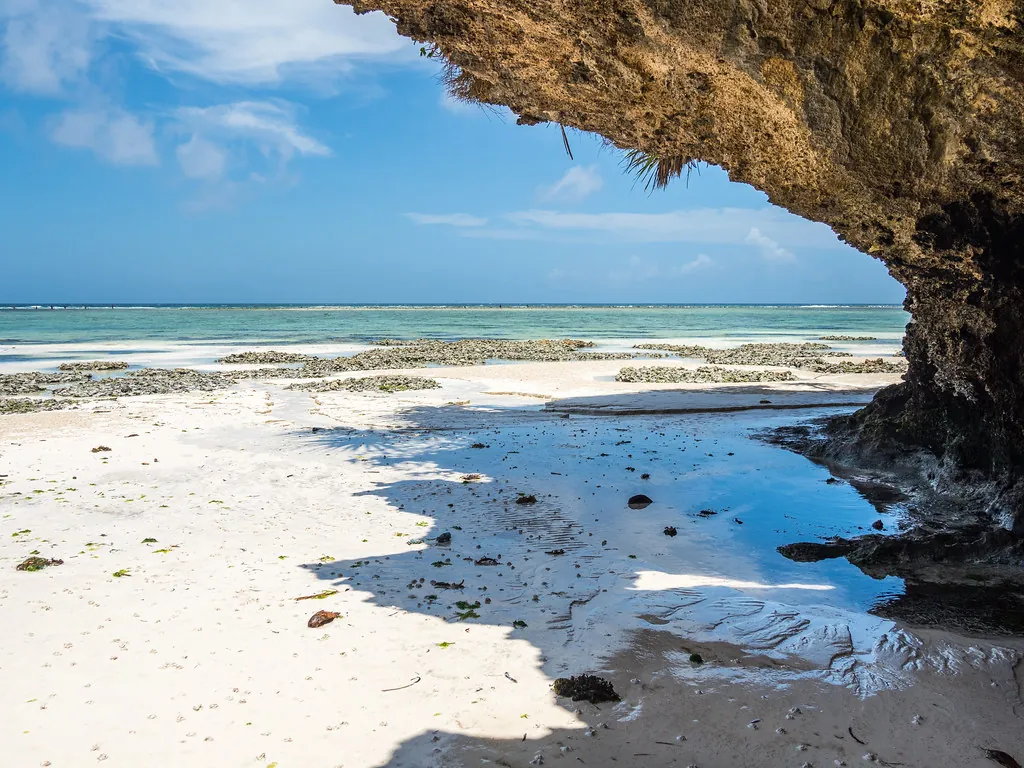 An overhanging rock ledge above white sand and shallow pools, the sea pulled back to a distant reef.