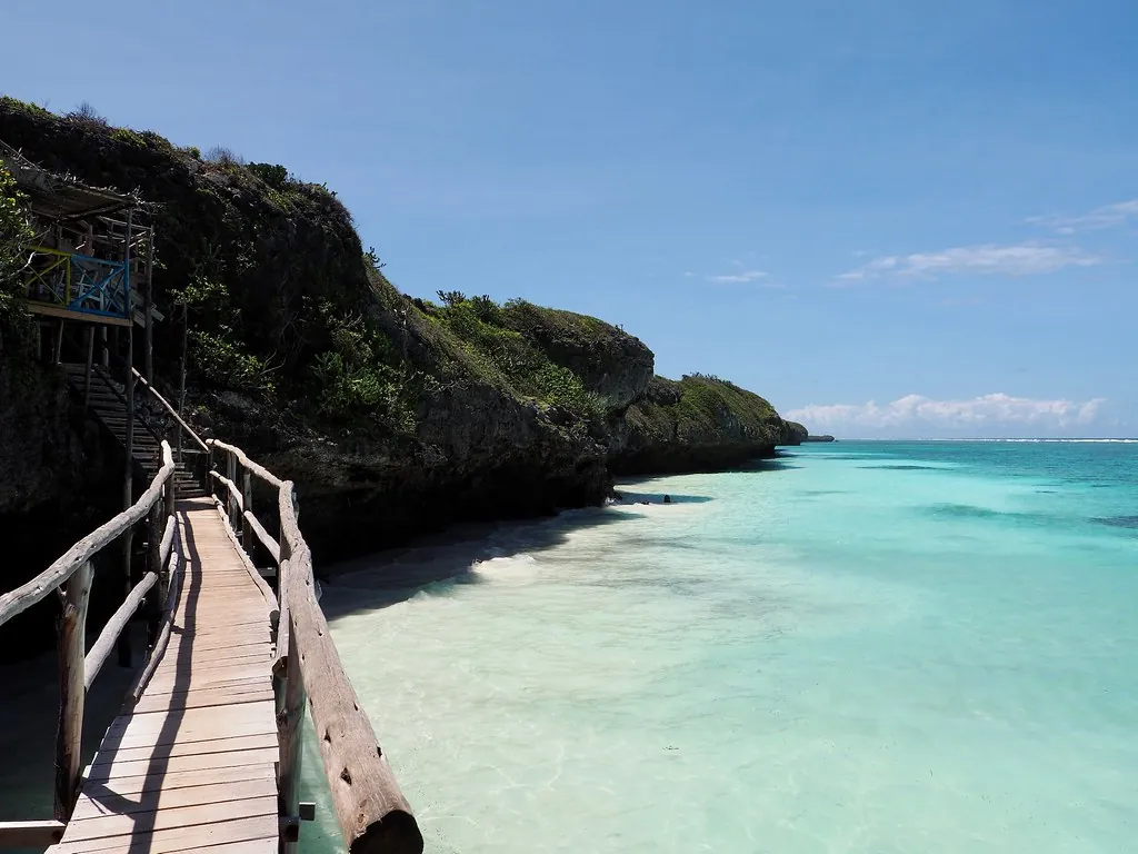 A wooden walkway along the foot of a vegetated cliff above a white beach and turquoise water.