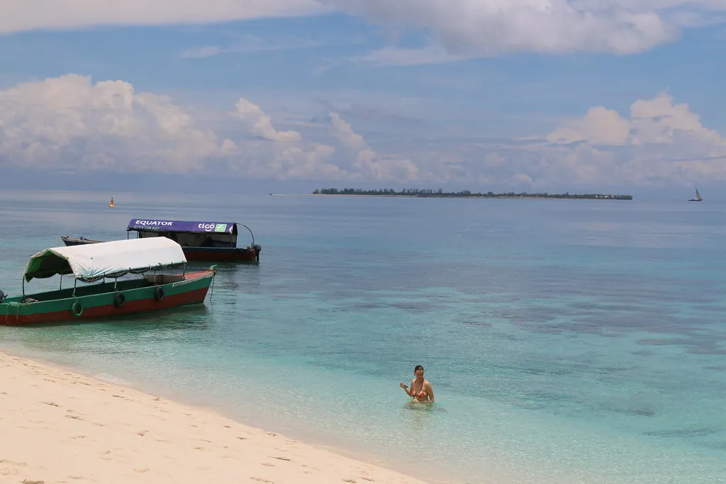 White sand sloping into clear turquoise water with a tour boat moored and a low island on the horizon