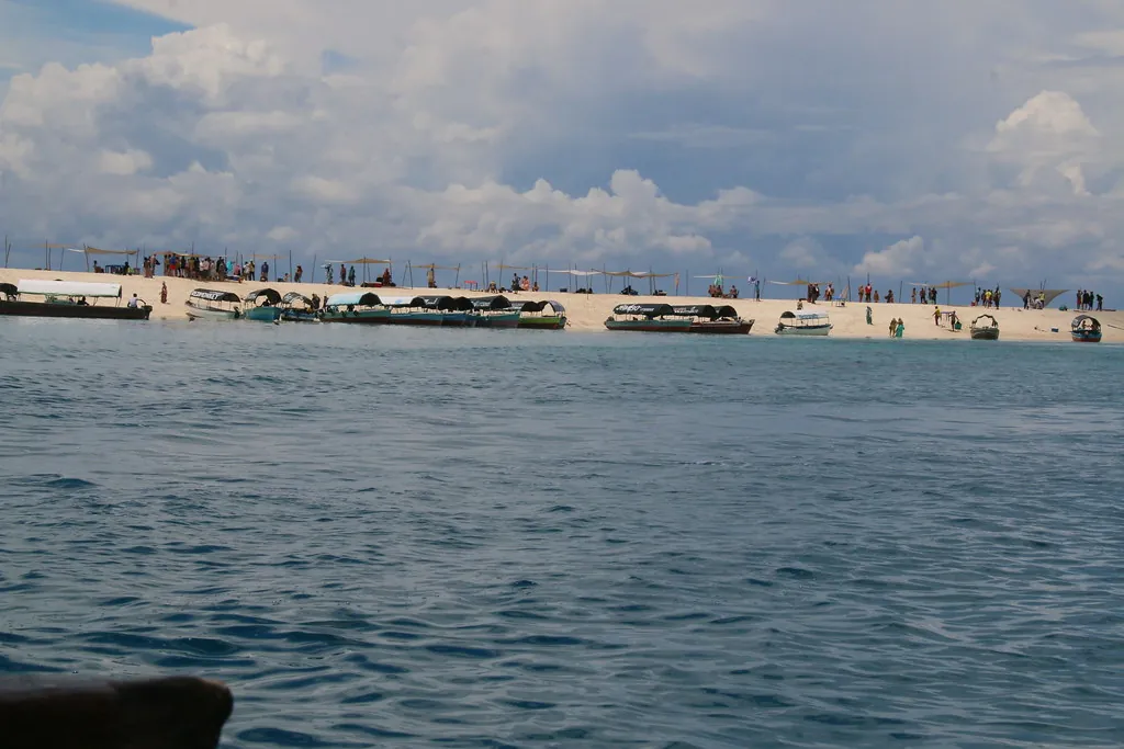A long, low white sandbank seen from a boat, lined with shade canopies, boats and people