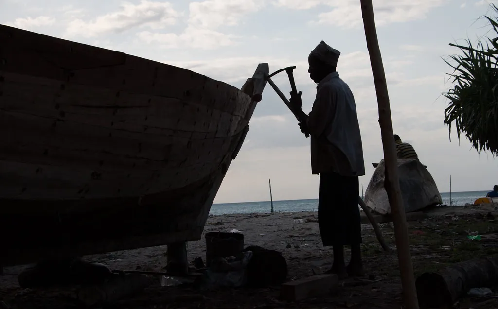 Silhouetted boatbuilder working with a hand tool on the hull of a wooden boat on the beach