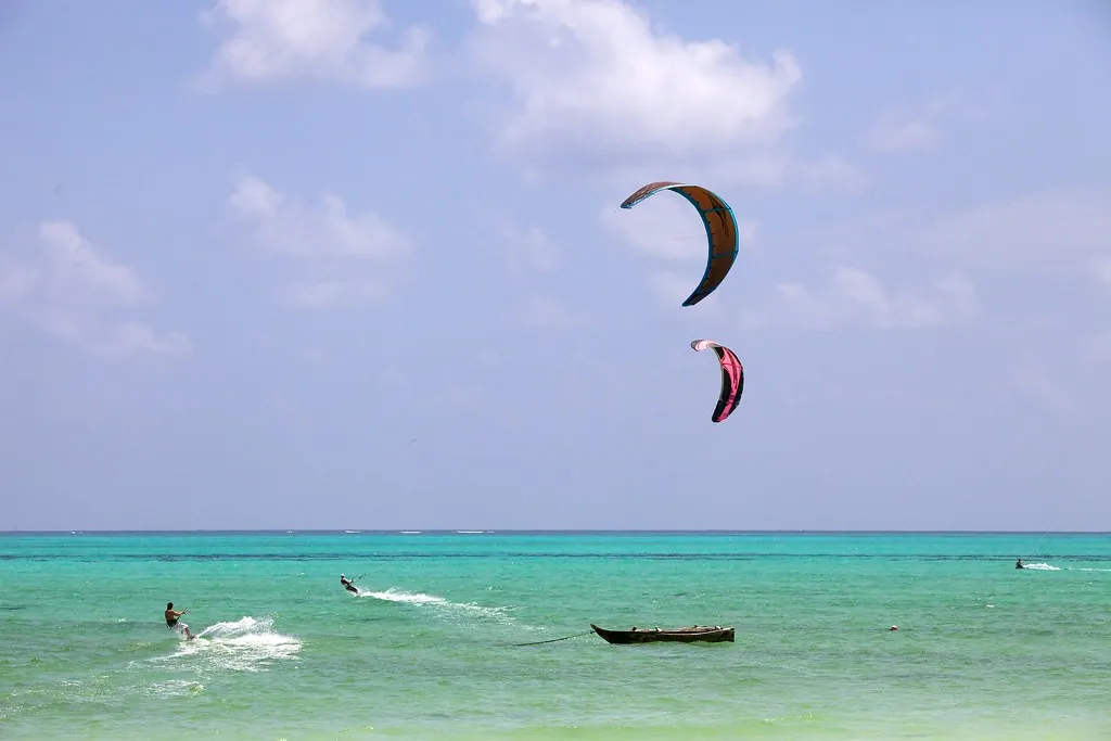 Two kitesurfers on bright turquoise water with a wooden outrigger boat moored nearby.