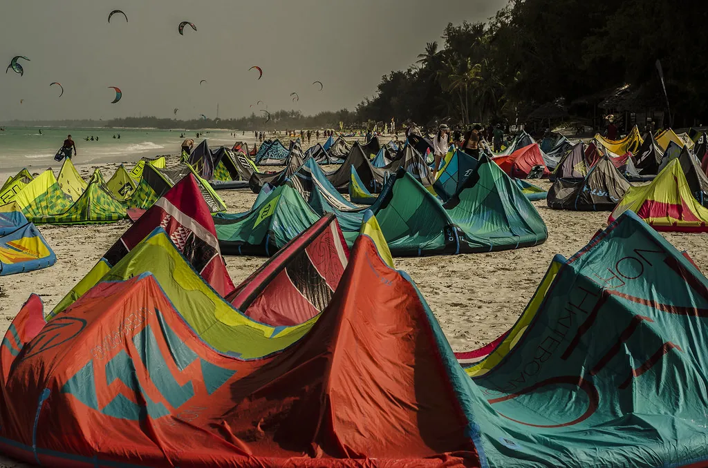 Dozens of colourful kitesurfing kites laid out on the sand with more kites flying over the sea.