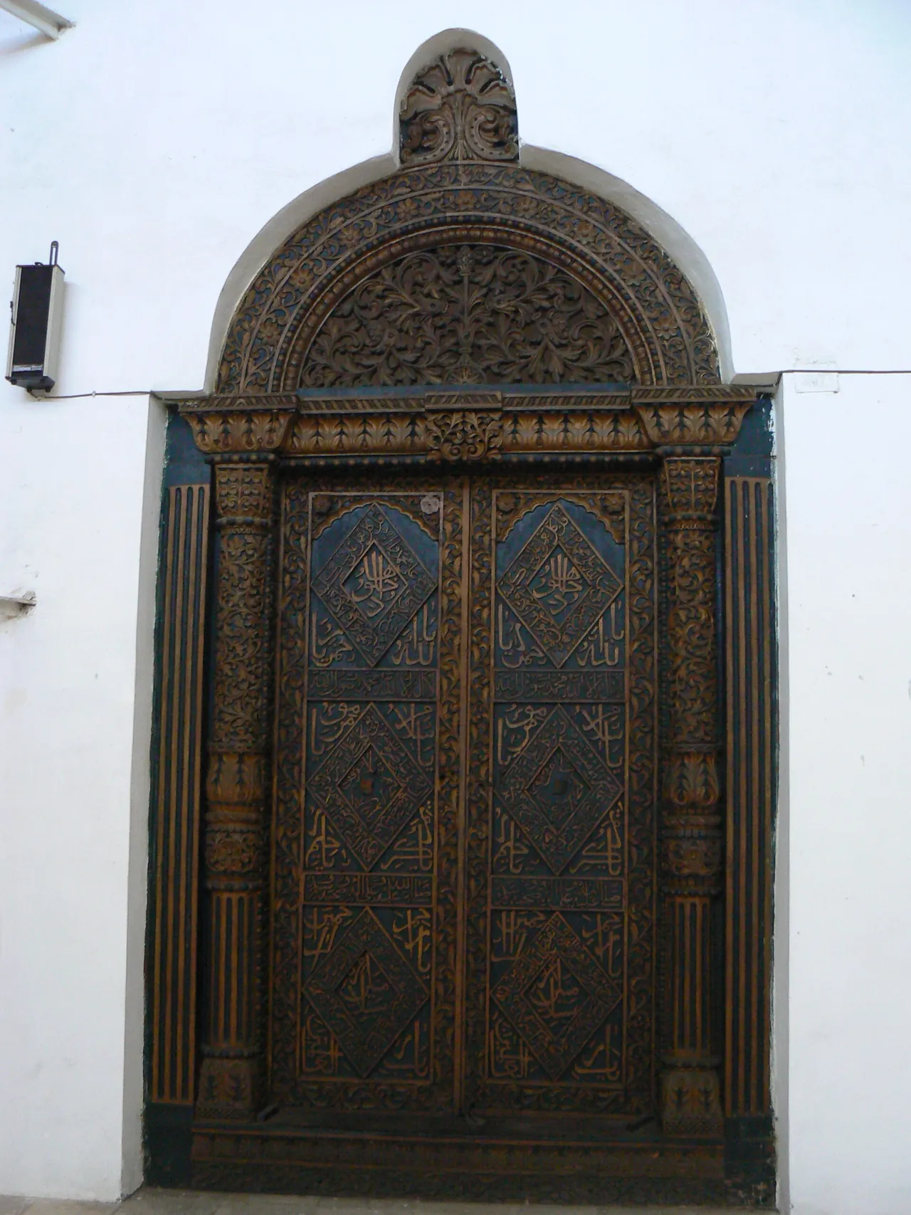 A tall carved wooden Zanzibari door with an arched, intricately carved frame set in a white wall