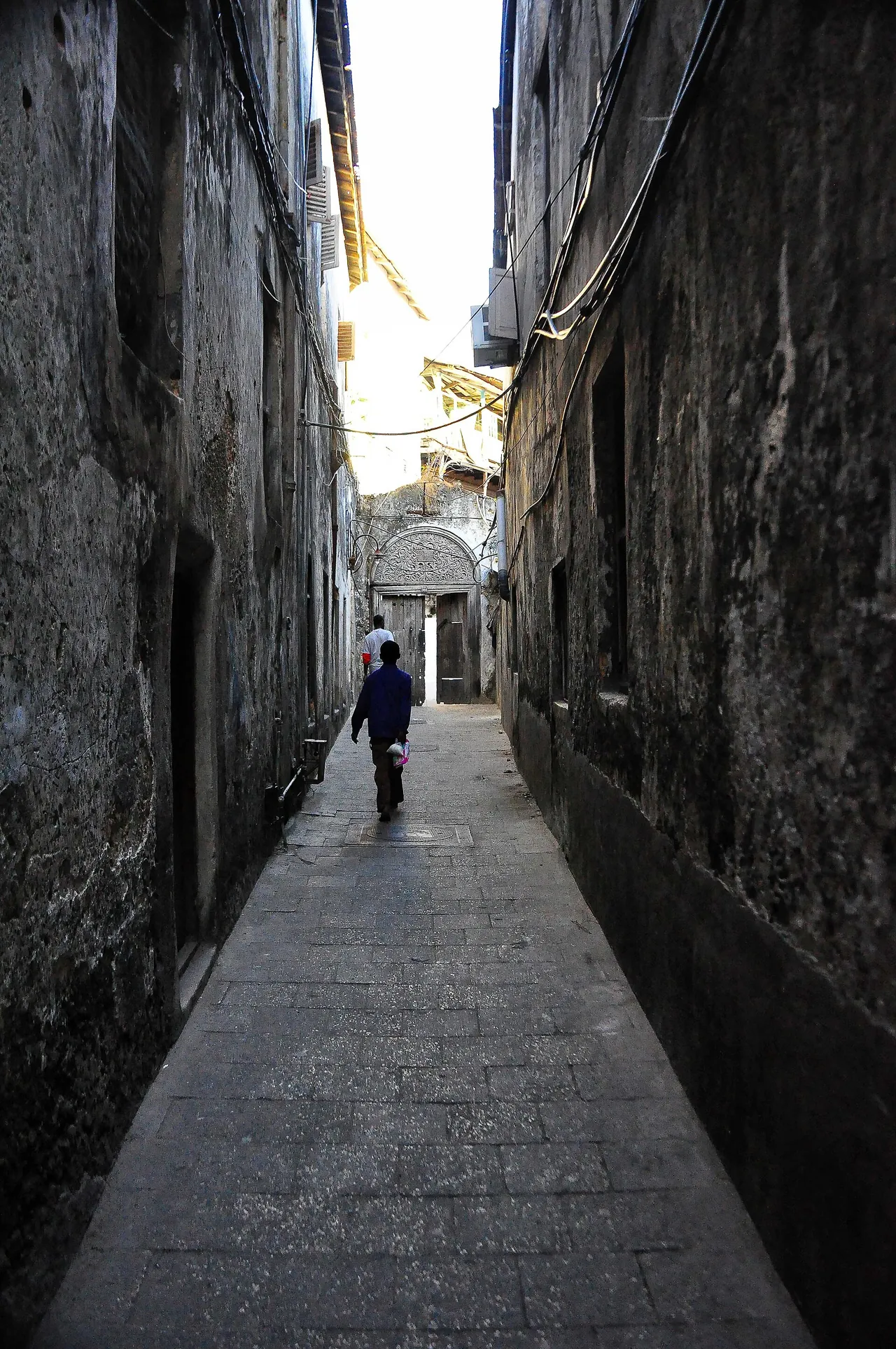 A long, shaded stone-paved alley between tall weathered coral-stone walls, with one person walking in the distance