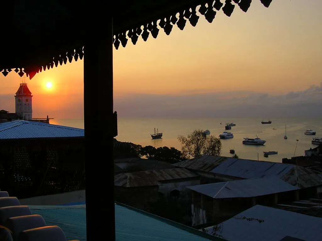 Sunset over the harbour and anchored boats, seen from under the carved eaves of a covered rooftop terrace, with tin roofs below
