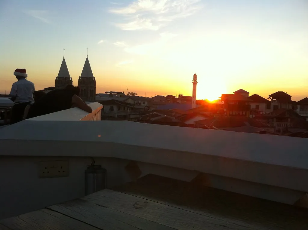 Sunset behind Stone Town's rooftops with the twin spires of St Joseph's Cathedral in silhouette and a person seated on a roof terrace