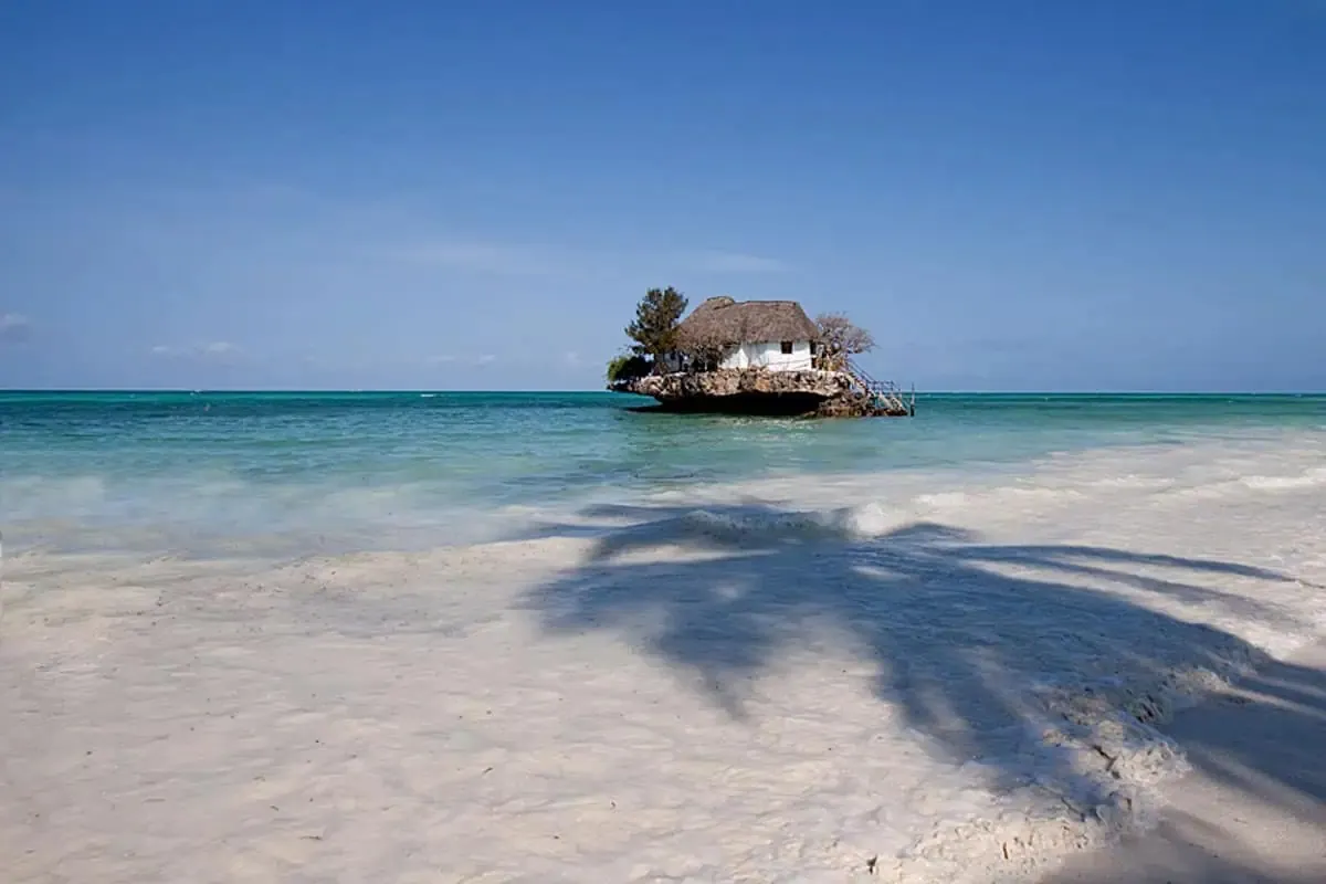 A small thatched restaurant on a rock offshore, seen from a white-sand beach with palm shadows and blue sea.