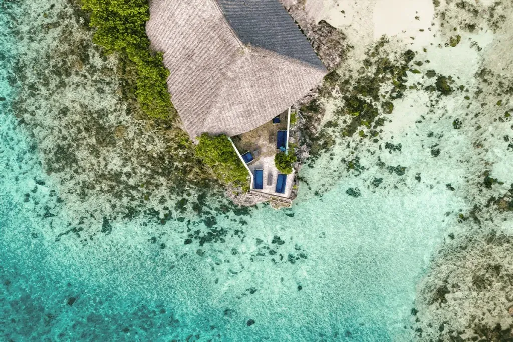 Aerial view looking straight down on a thatched building on a coral rock surrounded by turquoise shallows and reef.