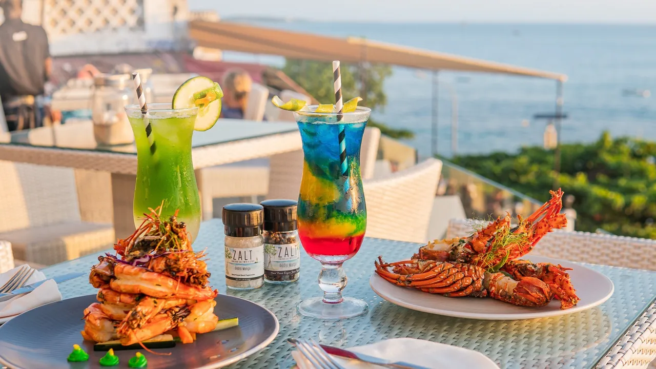 Grilled prawns, a lobster and two bright cocktails on a rooftop table with the sea behind