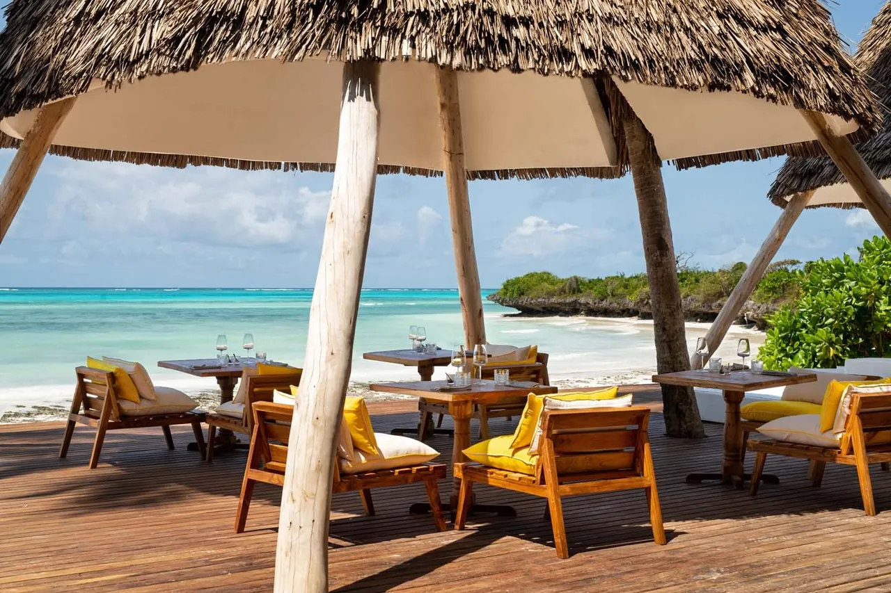 Wooden tables and armchairs with yellow cushions on a deck under a thatched parasol, facing a turquoise sea.