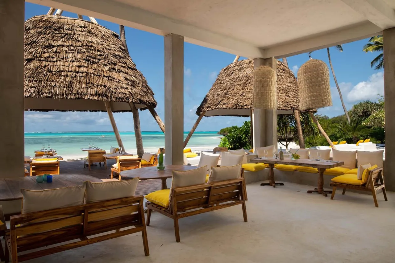 A covered lounge with sofas and low tables opening onto the beach and sea under thatched parasols.