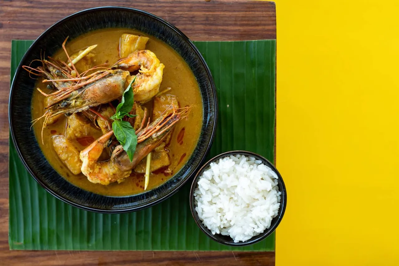 A prawn curry in a black bowl on a banana leaf with a bowl of white rice, against a yellow background.