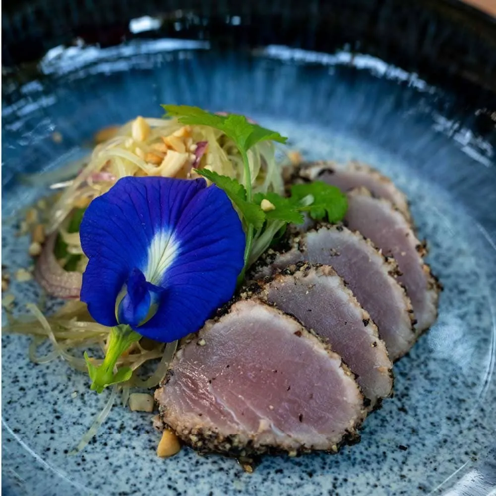 Slices of pink seared tuna with a blue flower garnish on a speckled blue plate.