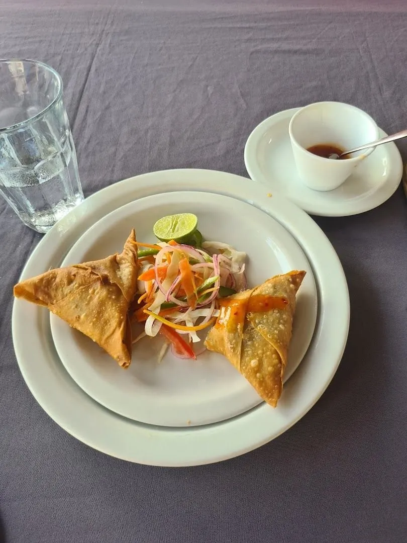 Two triangular samosas with a slaw garnish and a lime wedge on a white plate, a small cup of chili sauce beside it.