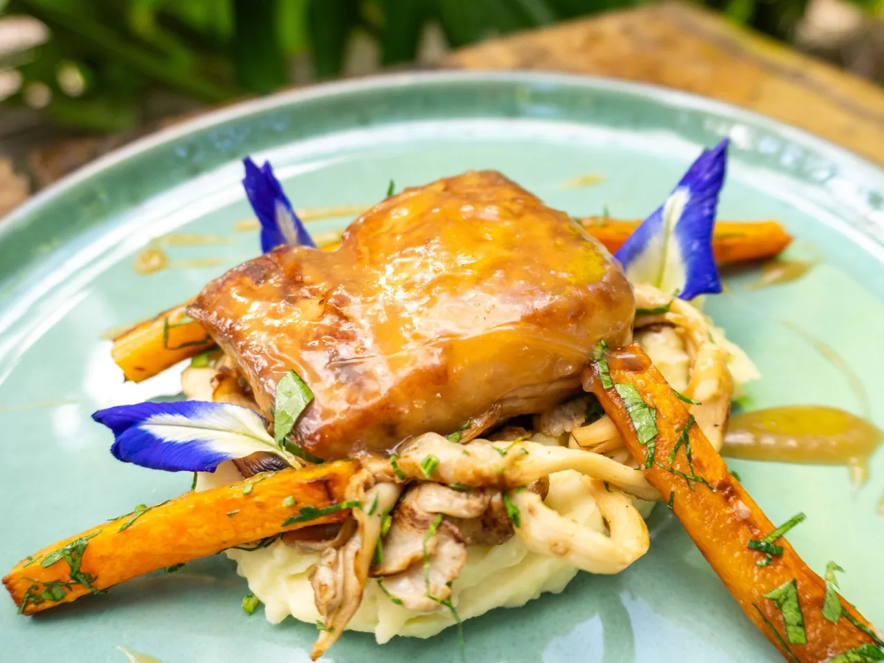 A glazed fillet of fish on mash with roasted carrots and blue butterfly-pea flowers, on a pale green plate.