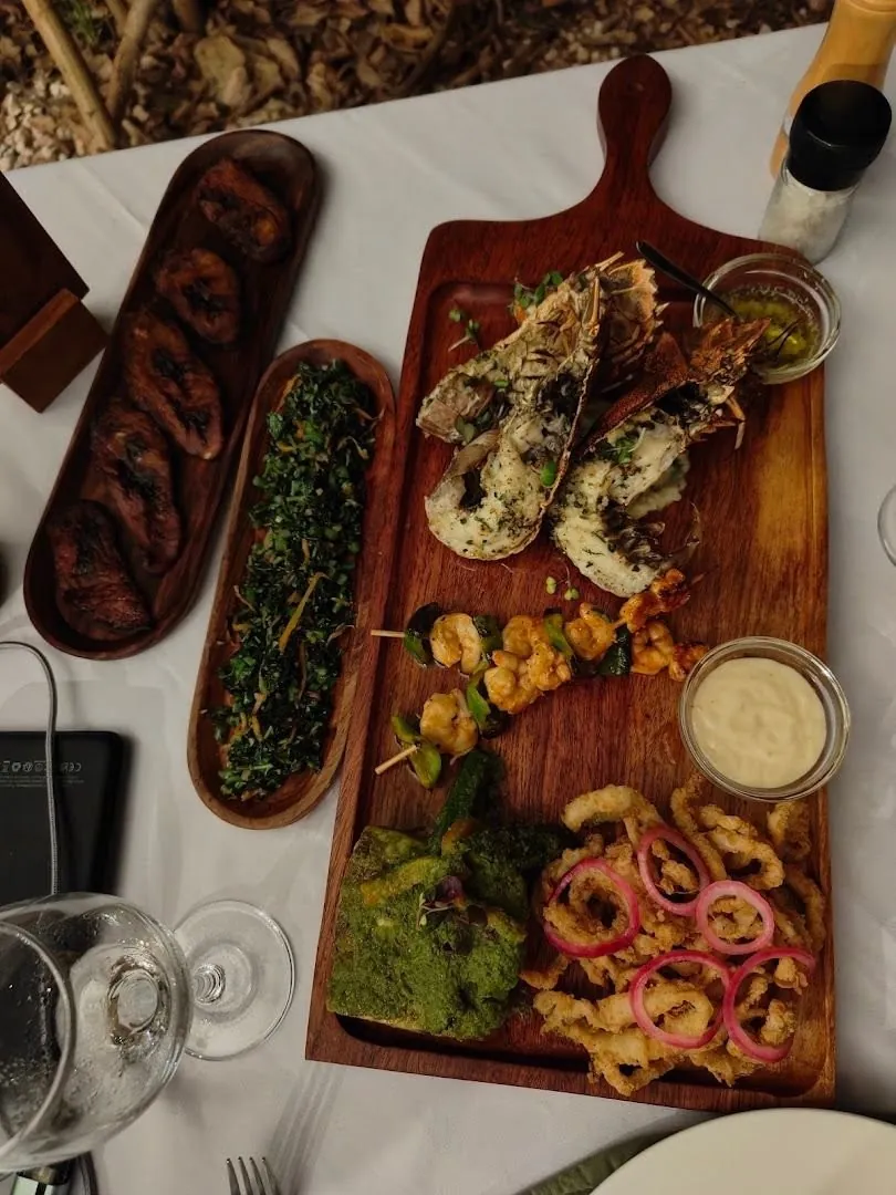 A wooden board of grilled lobster, prawn skewers and fried calamari rings, with side dishes of fried plantain and greens.