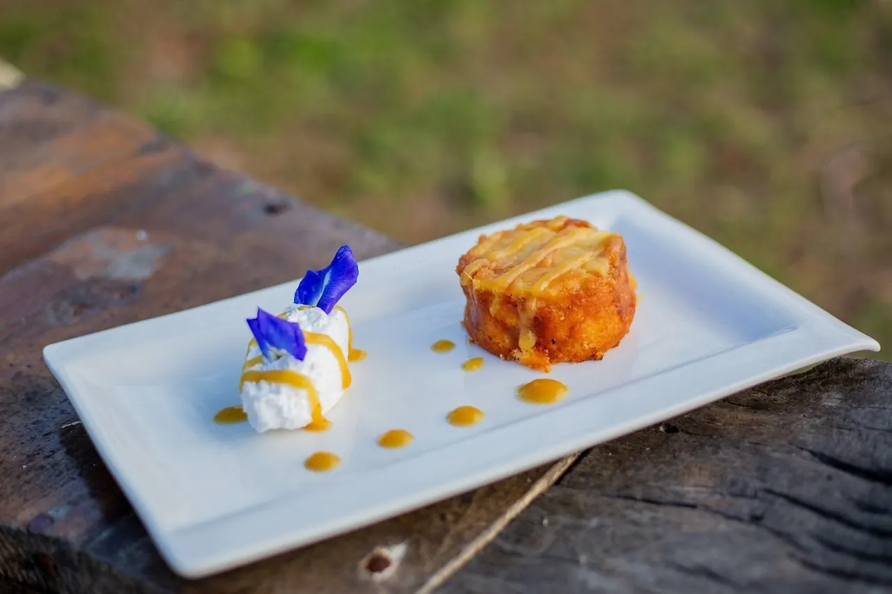 A small dessert plate with a round cake and a quenelle of cream topped with a blue flower, on a rustic wooden table.
