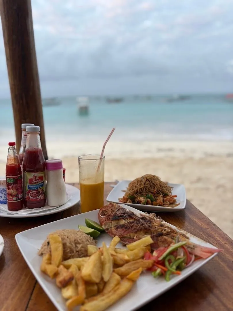 Grilled fish, noodles and a glass of juice on a table on the sand, with the sea and boats behind