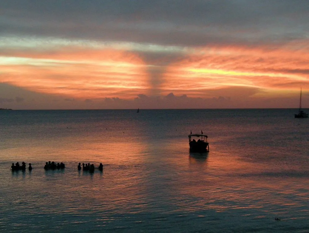 Silhouetted fishermen standing in the sea beside a boat under an orange sunset sky