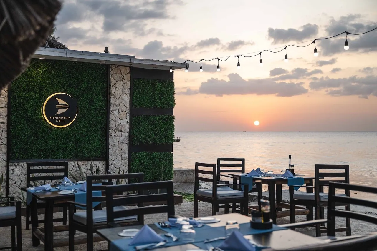 Outdoor tables on the sand under string lights, with the Fisherman's Grill sign on a green wall and the sun setting over the sea