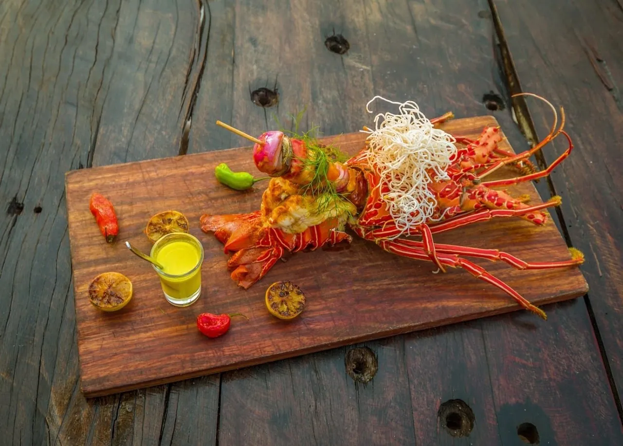 A grilled spiny lobster served on a wooden board with grilled lemon, chillies and a small glass of sauce
