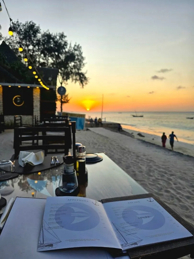 A table on Nungwi beach at sunset, with the restaurant's thatched building and lit sign to the left
