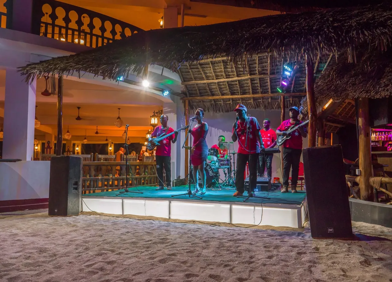 A band performing on a lit stage set on the sand at night, in front of a thatched building