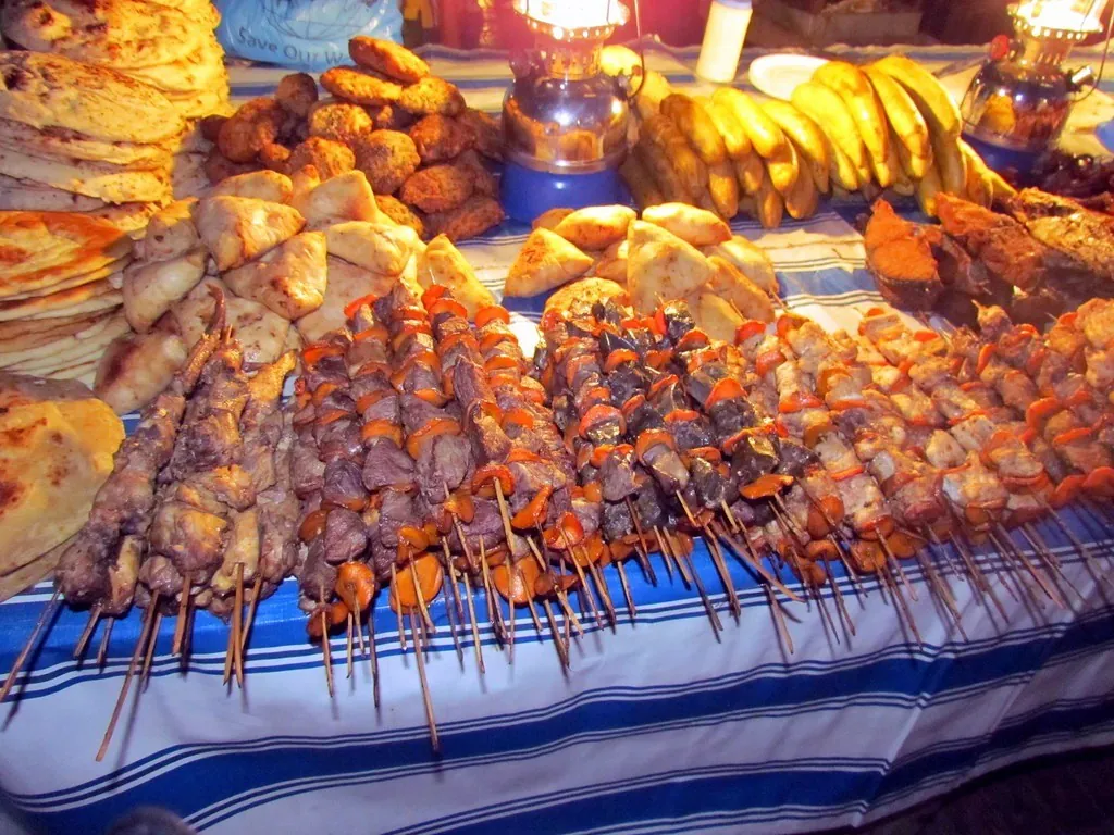 A stall table piled with skewers of grilled meat and seafood, chapatis, samosas and bananas
