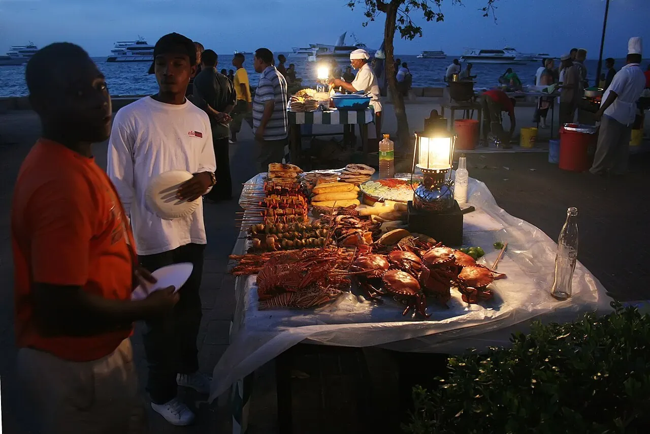 A seafront stall at dusk displaying lobsters, crabs and skewers beside a lantern, with the sea behind
