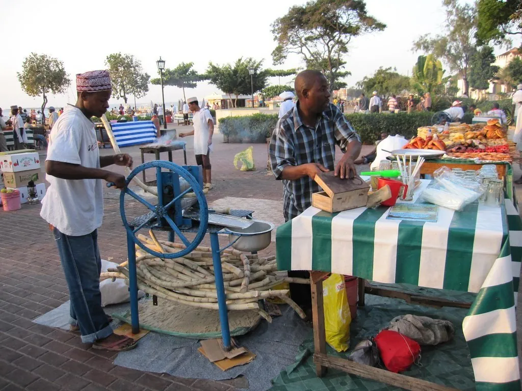 A vendor turning a hand-cranked press to squeeze sugarcane, with stalls being set up behind