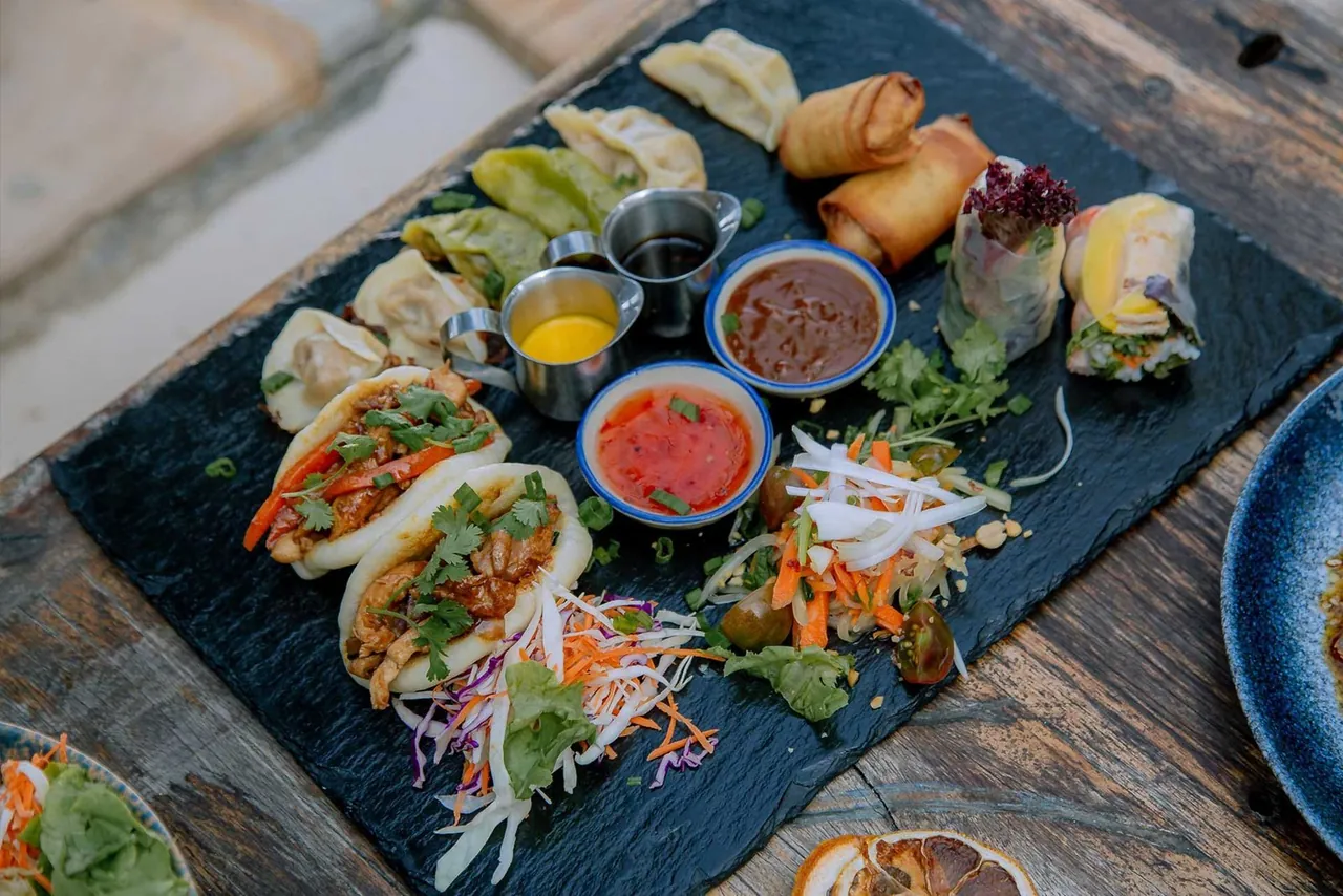 A slate board of dumplings, bao buns, spring rolls and summer rolls with dipping sauces
