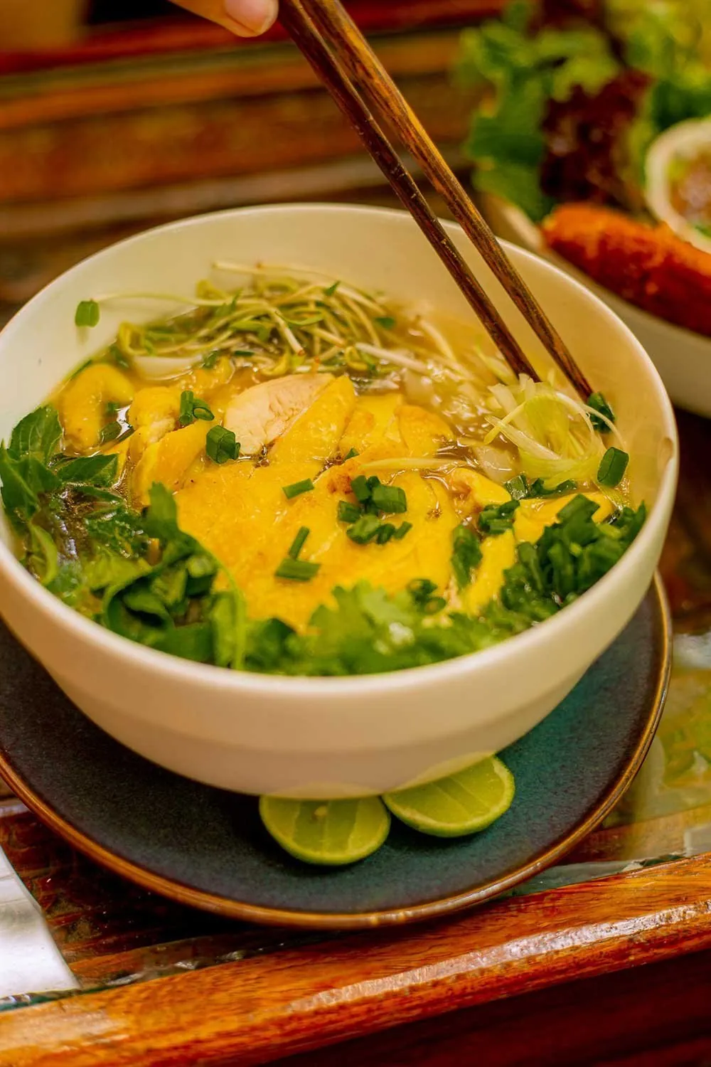 A bowl of chicken noodle soup with herbs and bean sprouts, chopsticks lifting noodles
