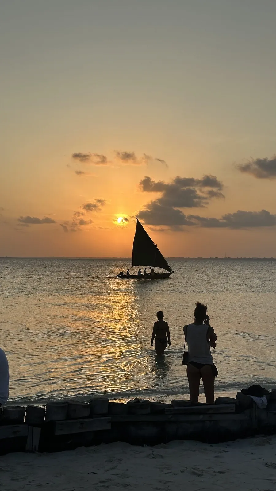 A dhow under sail silhouetted against an orange sunset, with people standing in the shallows in the foreground.