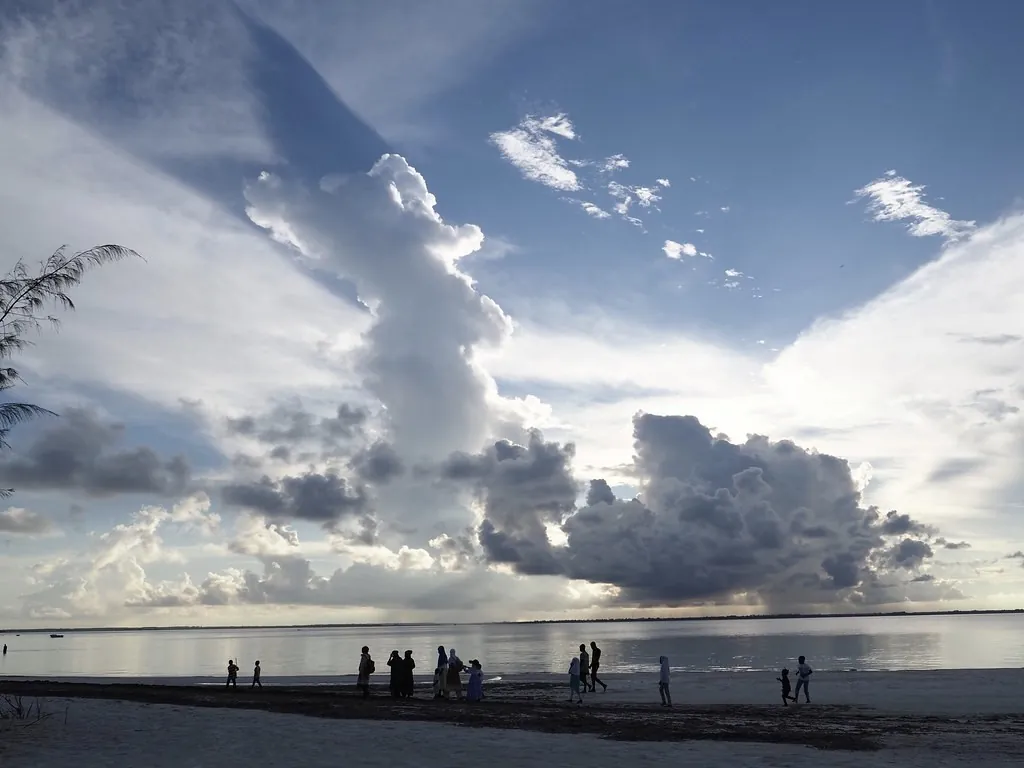 Towering clouds over a calm bay, with small figures standing on the shoreline.