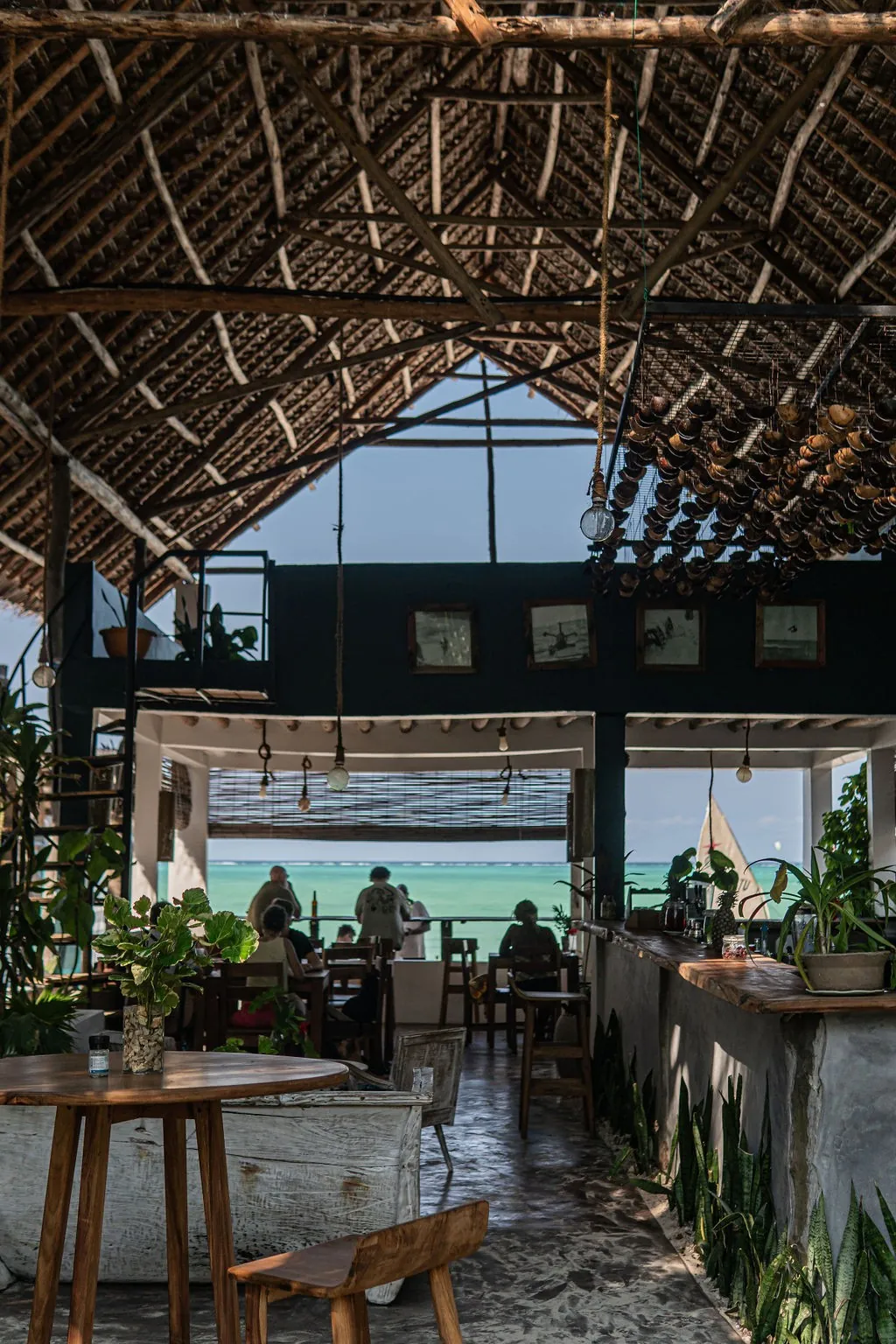 An open-sided café under a high thatched roof, with tables and plants and the turquoise sea visible beyond.
